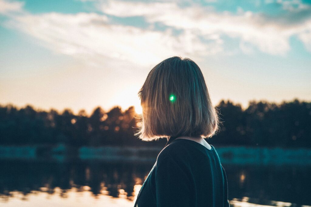 Sol og akne - på godt og ondt Alba. Silhuet af en person set fra bagsiden ved solnedgang ved en sø, med gyldne reflekser i vandet og blå himmel i baggrunden.